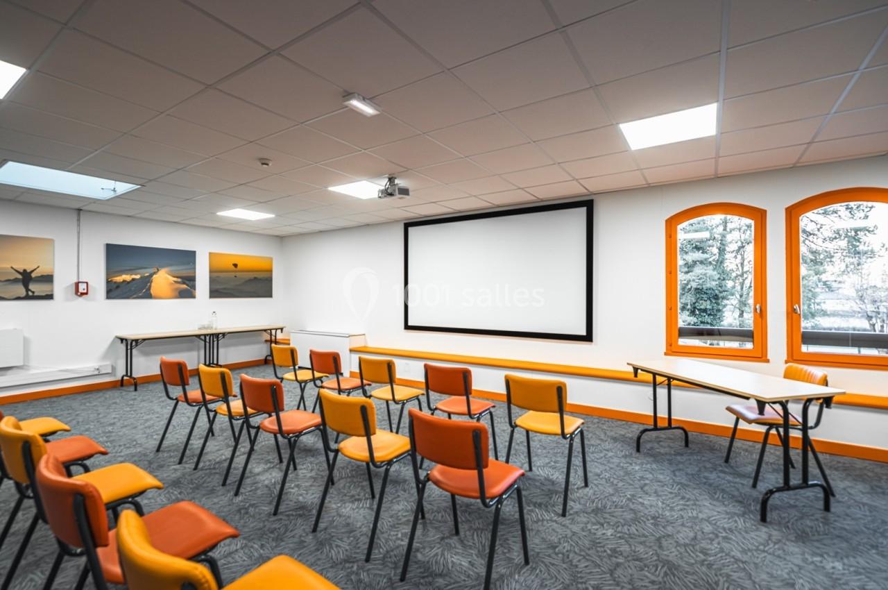 Salle de réunion lumineuse avec chaises orange, écran de projection, tableaux muraux et fenêtres donnant sur l'extérieur.