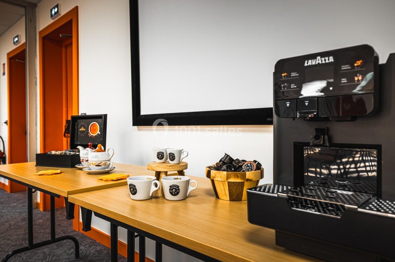 Machine à café automatique avec tasses, capsules et collations disposées sur une table en bois dans une salle lumineuse.