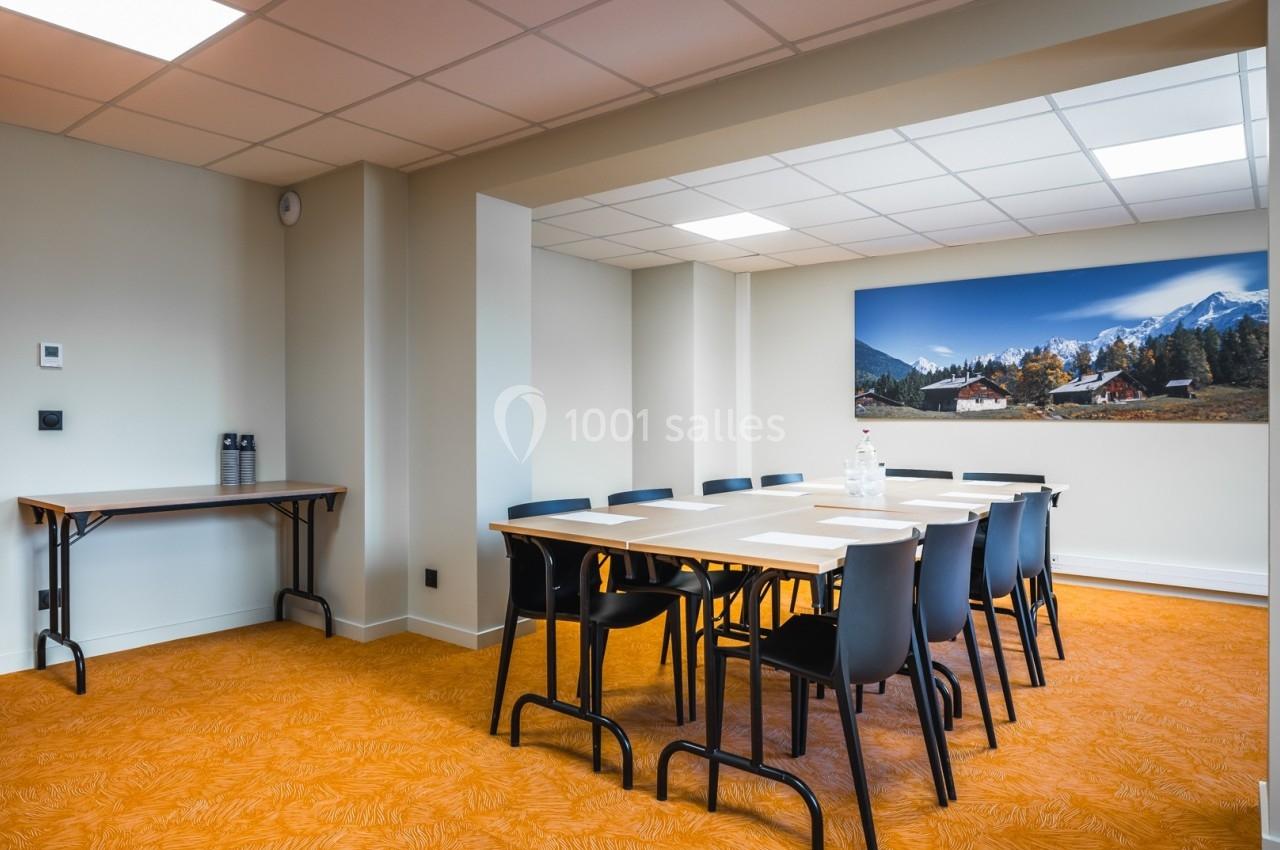 Salle de réunion lumineuse avec table rectangulaire, chaises noires, sol orange et tableau mural représentant un paysage…