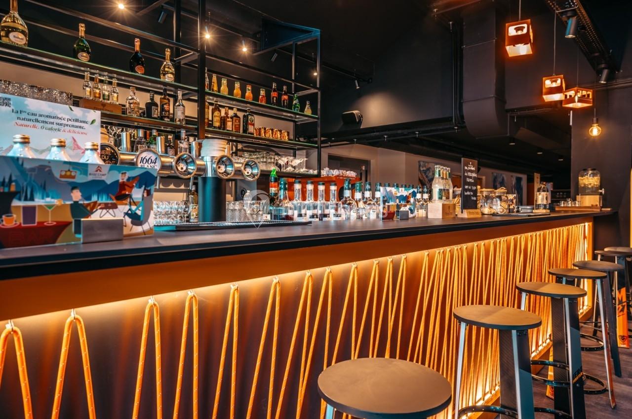 Bar moderne avec comptoir éclairé par des lumières LED, tabourets hauts et étagères garnies de bouteilles et verres.