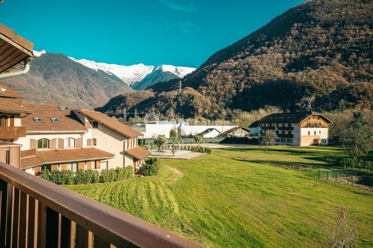 Vue depuis un balcon sur un paysage de montagne avec des maisons, des prairies et des sommets enneigés sous un ciel bleu.