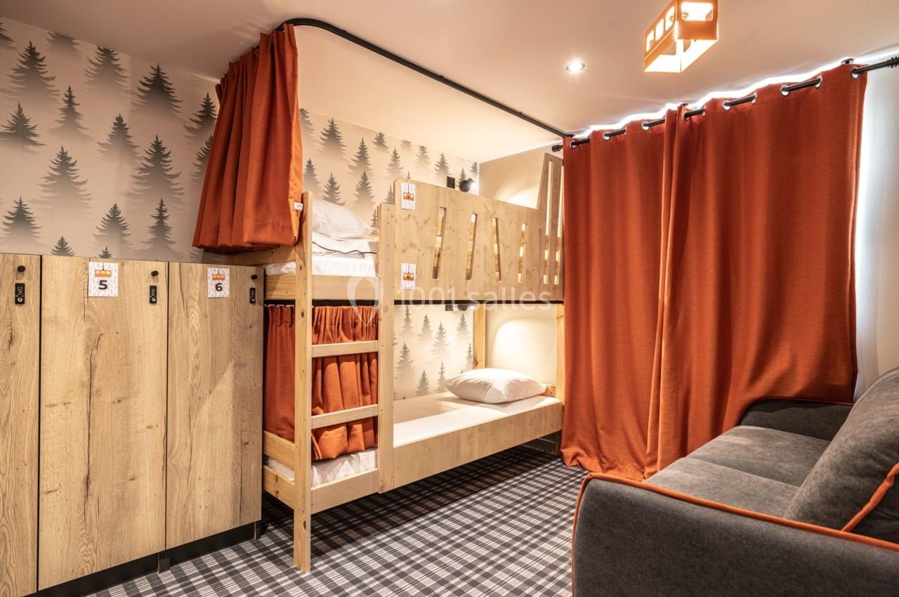 Chambre avec lits superposés en bois, rideaux orange, casiers en bois et canapé sur un sol à motif carreaux.