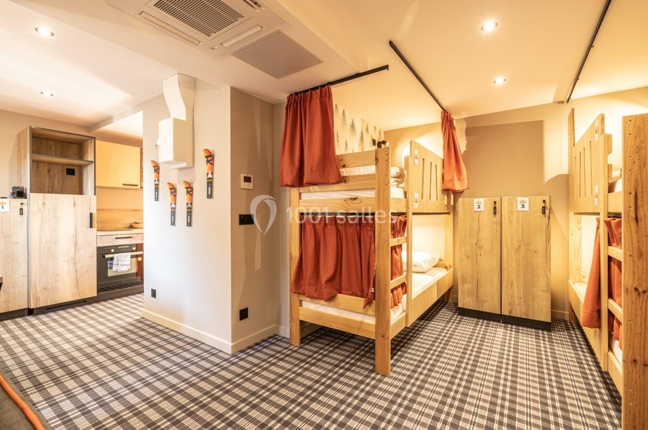 Dortoir avec lits superposés en bois, rideaux pour l'intimité, casiers individuels et coin cuisine en arrière-plan.