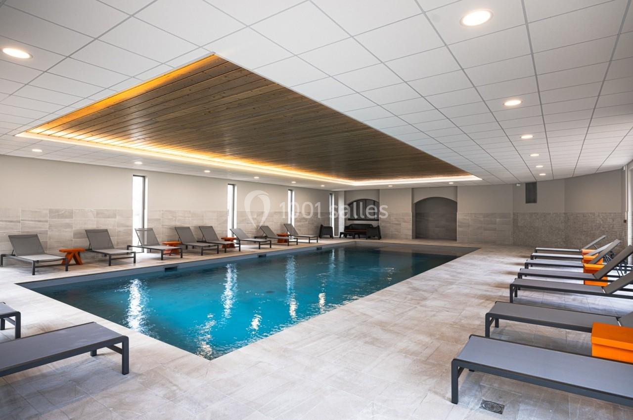 Piscine intérieure entourée de chaises longues grises avec coussins orange, éclairage moderne au plafond.