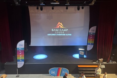 Un groupe de musiciens joue sur scène devant un écran affichant le logo et le slogan ’Base Camp Lodge’.