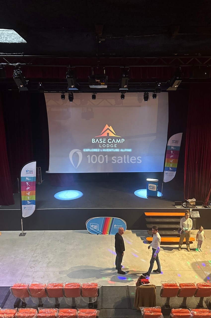 Vue d'une scène avec un écran projetant le logo ’Base Camp Lodge’, entourée de rideaux rouges et de personnes discutant.