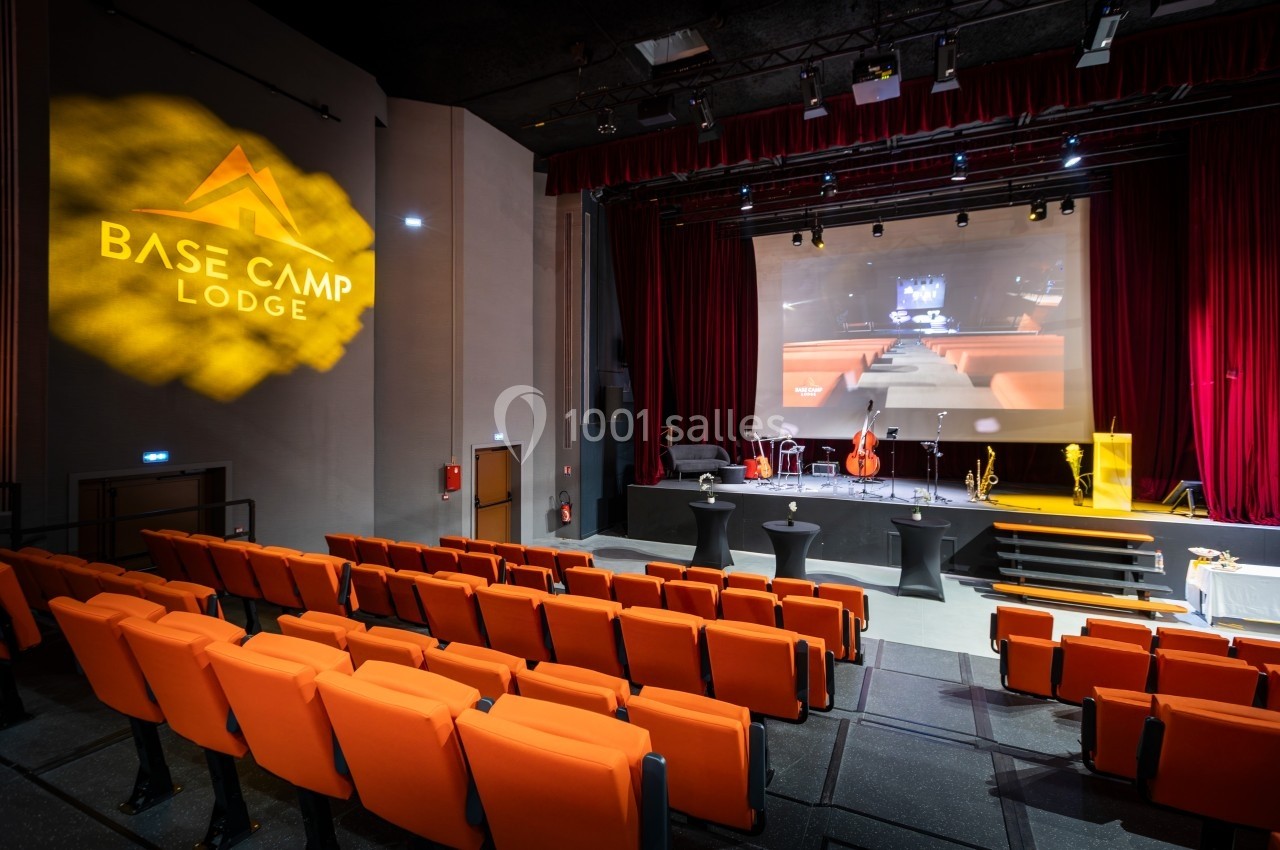 Salle de conférence avec sièges orange, scène équipée d'instruments de musique et écran de projection allumé.