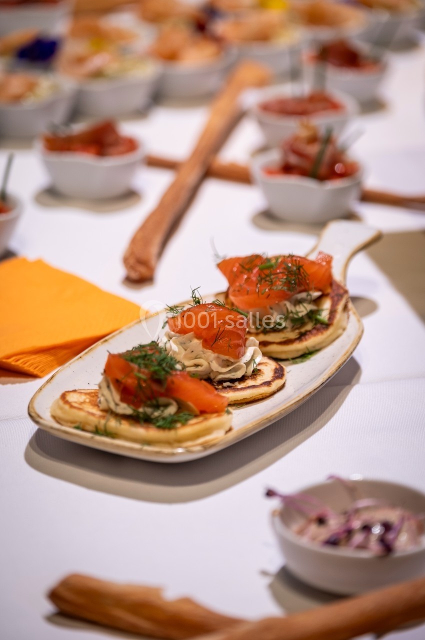Assiette de blinis garnis de saumon fumé et crème, entourée de petites coupelles de condiments sur une table dressée.