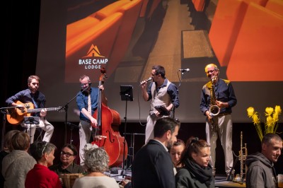 Un groupe de musiciens joue sur scène devant un écran affichant le logo et le slogan ’Base Camp Lodge’.