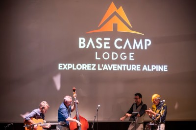 Un groupe de musiciens joue sur scène devant un écran affichant le logo et le slogan ’Base Camp Lodge’.