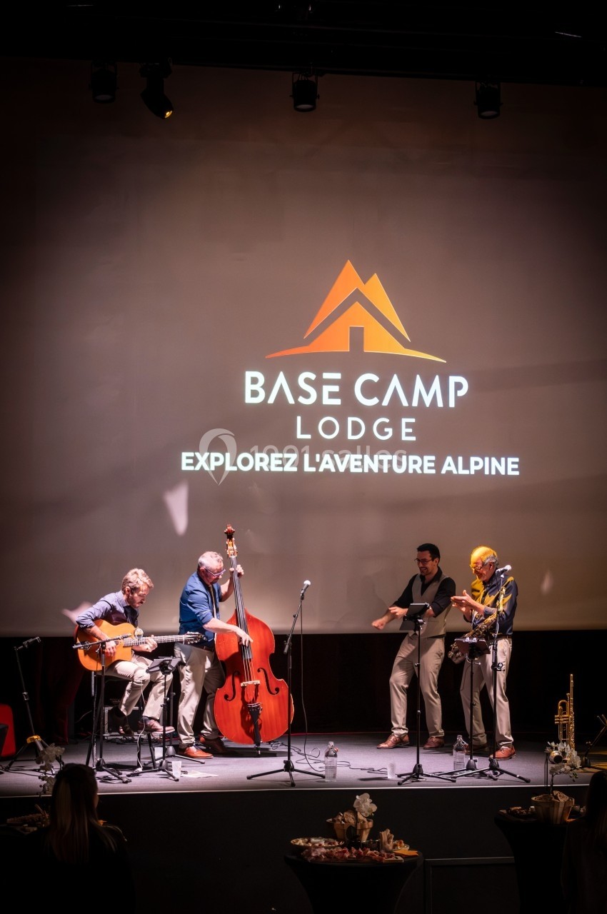 Un groupe de musiciens joue sur scène devant un écran affichant le logo et le slogan ’Base Camp Lodge’.