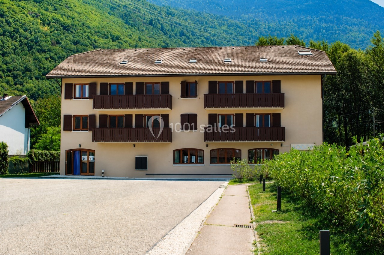 Bâtiment résidentiel à trois étages avec balcons en bois, entouré de verdure et de montagnes en arrière-plan.