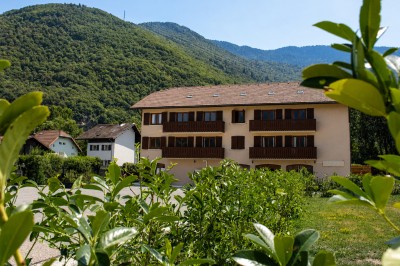 Bâtiment résidentiel à trois étages avec balcons en bois, entouré de verdure et de montagnes en arrière-plan.