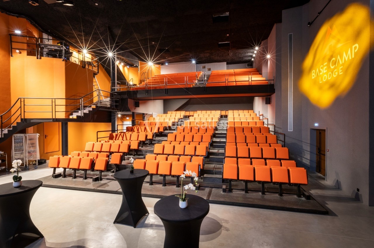 Salle de conférence moderne avec des sièges orange, un éclairage tamisé et des tables hautes au premier plan.