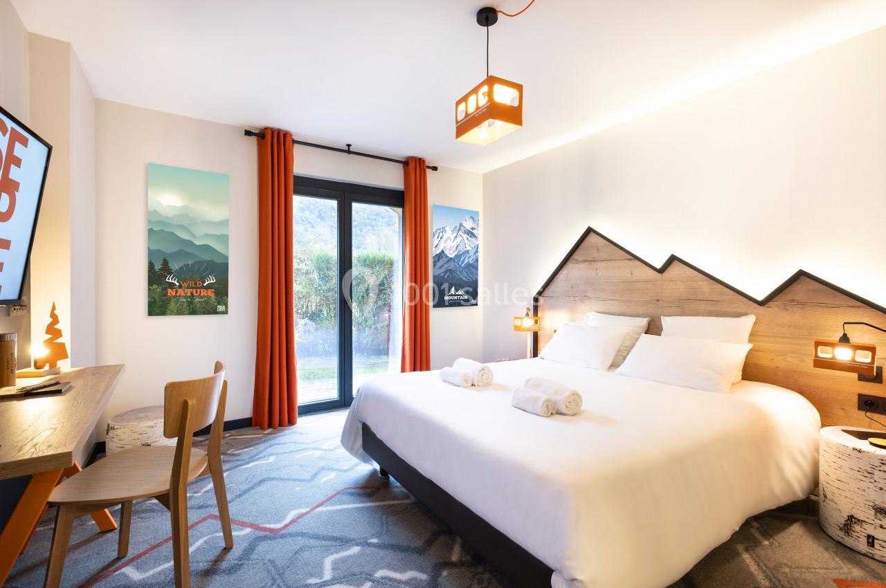 Chambre lumineuse avec un lit double, bureau, télévision, grandes fenêtres et vue sur un jardin verdoyant.