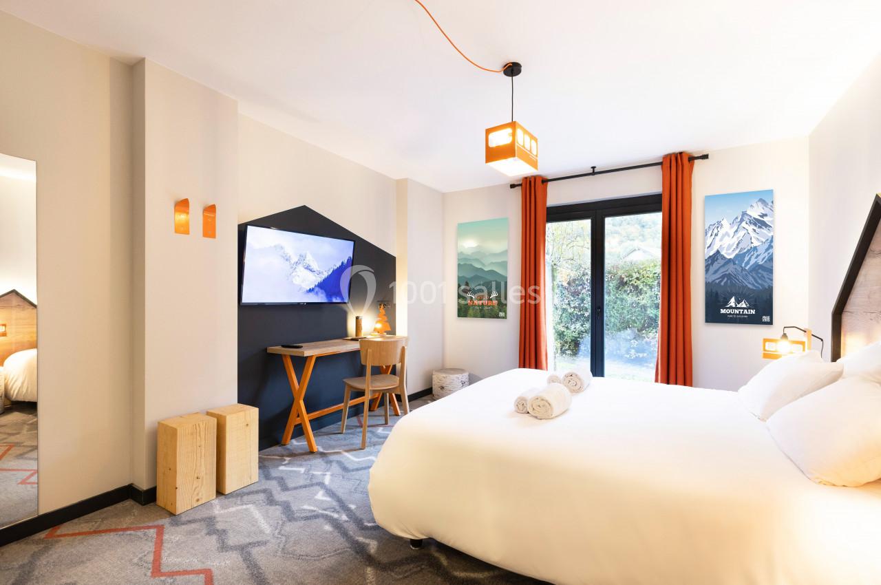 Chambre lumineuse avec lit double, bureau, télévision, décorations murales et grande fenêtre donnant sur un jardin.