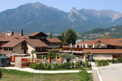 Bâtiment résidentiel à trois étages avec balcons en bois, entouré de verdure et de montagnes en arrière-plan.
