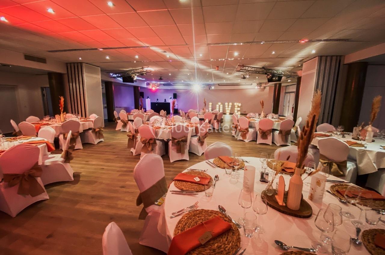 Salle de réception décorée avec des tables rondes, nappes blanches, chaises ornées de rubans et éclairage chaleureux.