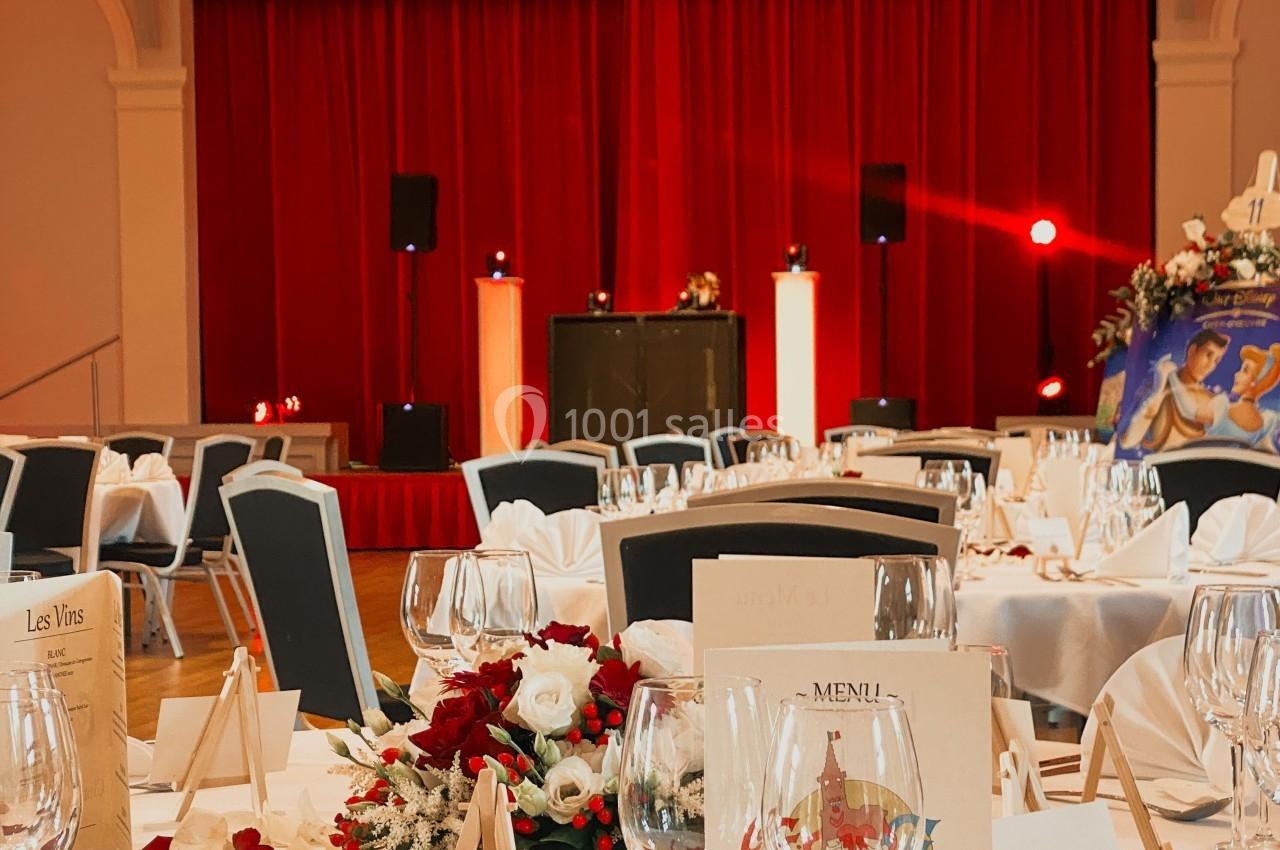 Salle de réception décorée avec des tables dressées, des fleurs et une scène avec rideaux rouges en arrière-plan.