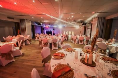 Salle de réception décorée pour un événement, avec tables dressées, chaises blanches et éclairage chaleureux.