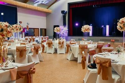 Salle de réception décorée avec des tables rondes, nappes blanches, chaises ornées de nœuds orange et centres de table…
