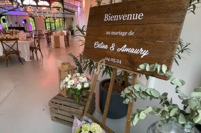 Panneau en bois décoré de fleurs, accueillant les invités à un mariage dans une salle lumineuse et aménagée.