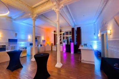 Salle lumineuse avec colonnes blanches, parquet en bois, tables hautes noires et éclairages colorés.