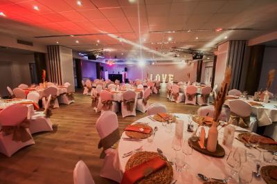 Salle de réception élégante avec tables rondes dressées, décorations florales et lumière naturelle entrant par les fenêtres.