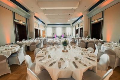 Salle de réception élégante avec tables rondes dressées, décorations florales et lumière naturelle entrant par les fenêtres.