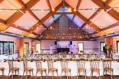 Salle de réception élégante avec tables rondes dressées, décorations florales et lumière naturelle entrant par les fenêtres.