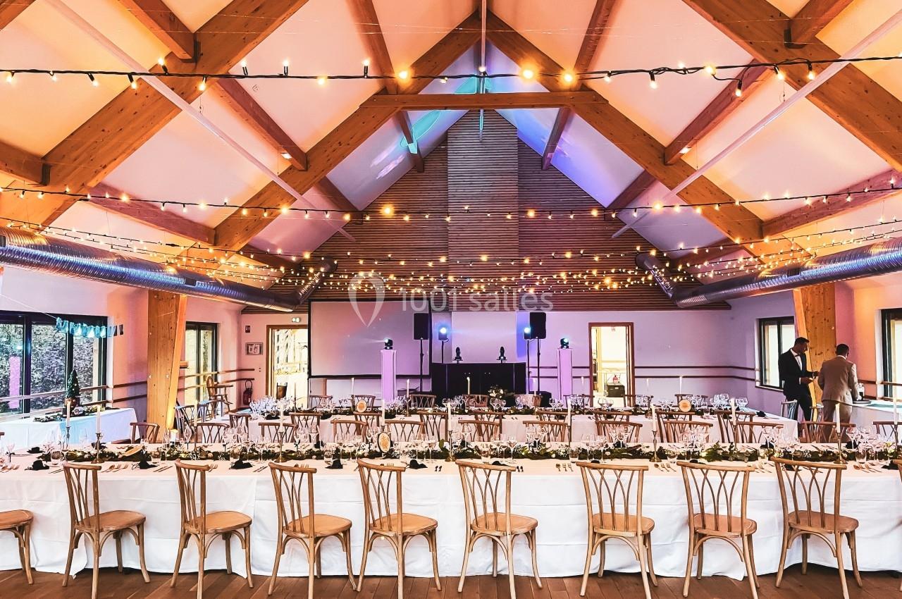 Grande salle décorée pour un événement, avec tables dressées, chaises en bois et guirlandes lumineuses suspendues.