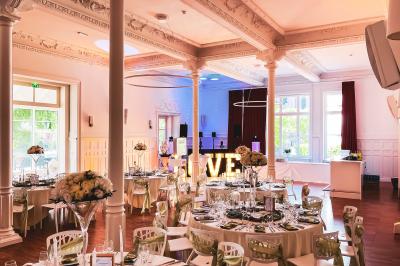 Salle de réception élégante avec tables rondes dressées, décorations florales et lumière naturelle entrant par les fenêtres.