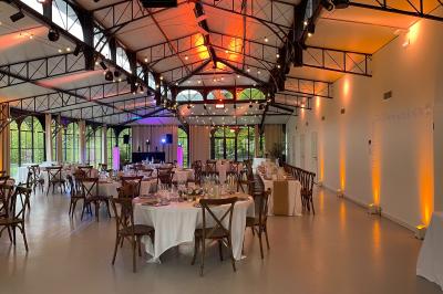 Salle de réception élégante avec tables rondes dressées, décorations florales et lumière naturelle entrant par les fenêtres.