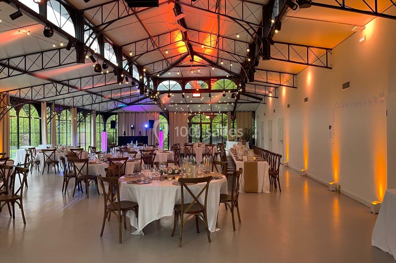 Salle de réception lumineuse avec tables rondes dressées, chaises en bois et éclairage chaleureux.