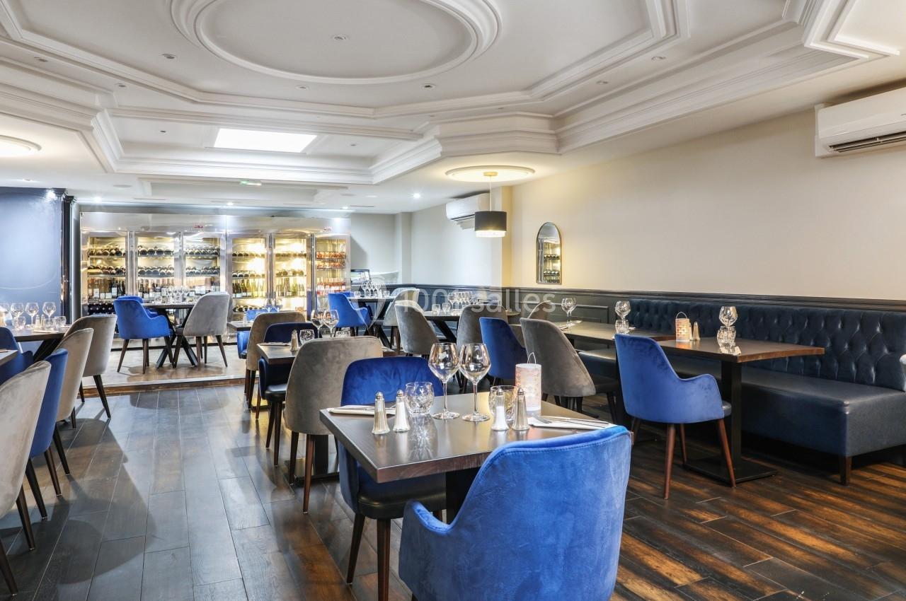 Salle de restaurant élégante avec tables dressées, chaises bleues et grises, parquet en bois et éclairage chaleureux.