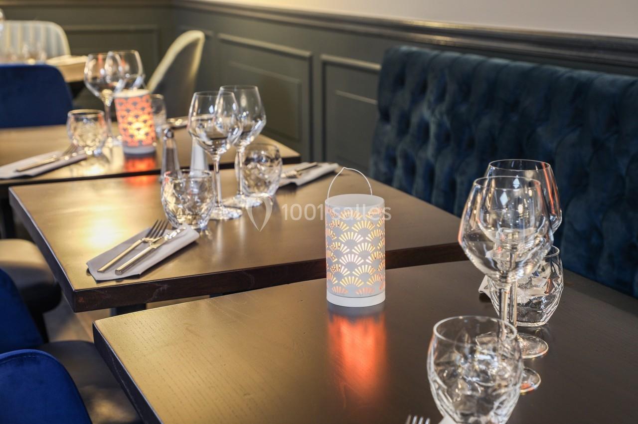 Tables de restaurant élégamment dressées avec verres, couverts et photophores lumineux sur des nappes sombres.