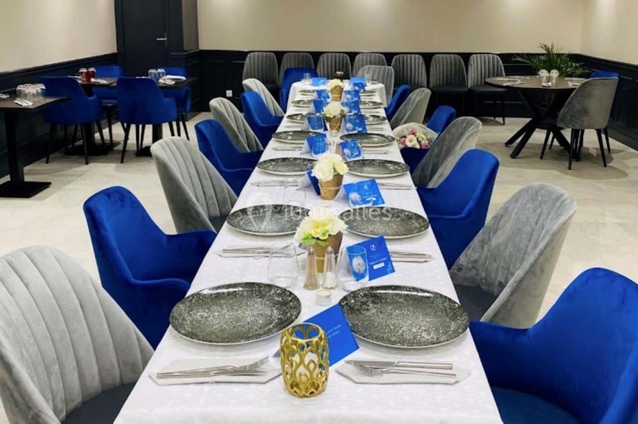 Table dressée avec nappes blanches, assiettes décoratives, fleurs et chaises bleues et grises dans une salle élégante.