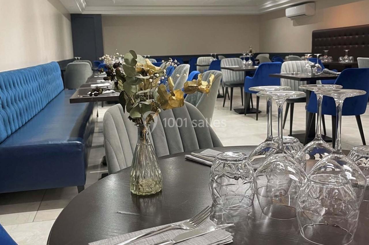 Salle de restaurant moderne avec tables dressées, chaises bleues et grises, et un vase de fleurs séchées au premier plan.
