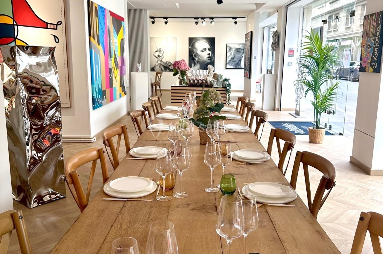 Grande table en bois dressée avec vaisselle et verres, entourée de chaises, dans une salle lumineuse décorée d'œuvres d'art.