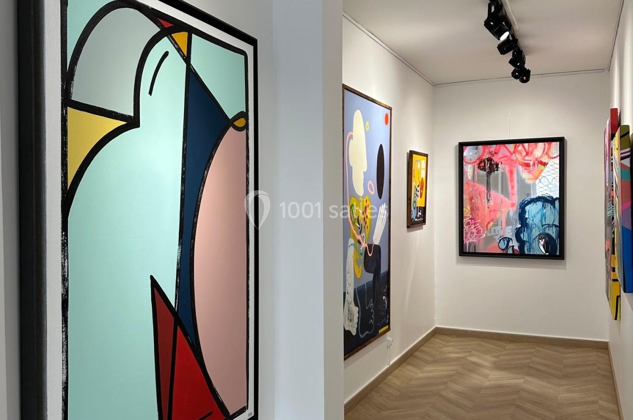 Vue d'une galerie d'art avec des tableaux modernes et colorés accrochés sur des murs blancs, éclairés par des spots.