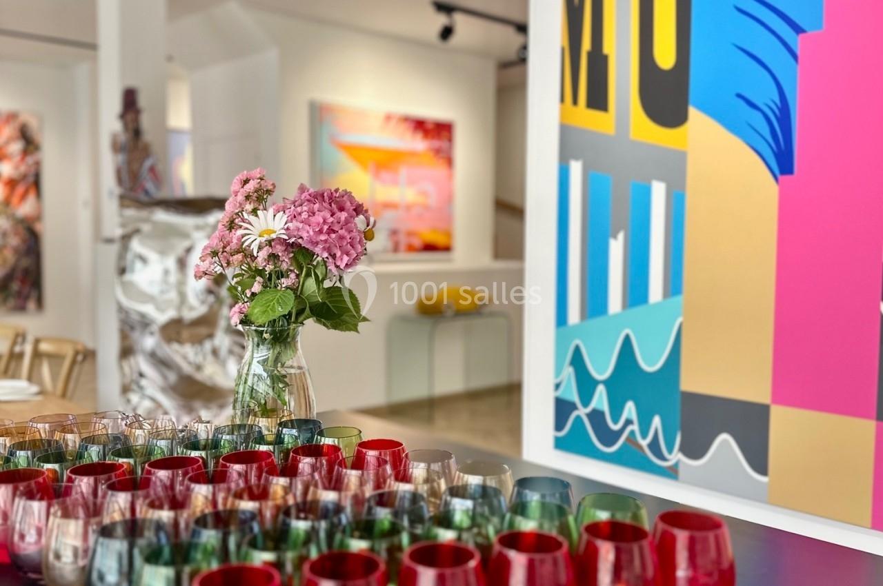Vase de fleurs roses sur une table noire entourée de verres colorés, dans une galerie d'art moderne lumineuse.