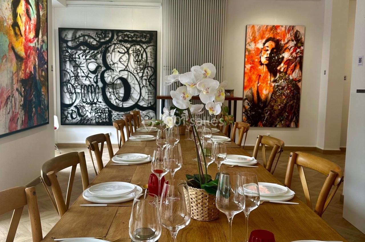 Table en bois dressée avec vaisselle et verres, entourée de chaises, dans une salle décorée de tableaux modernes.