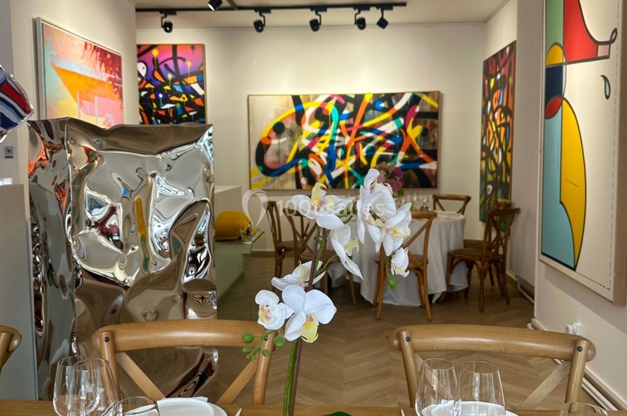 Salle d'exposition lumineuse avec des œuvres d'art colorées accrochées aux murs, table dressée au premier plan.