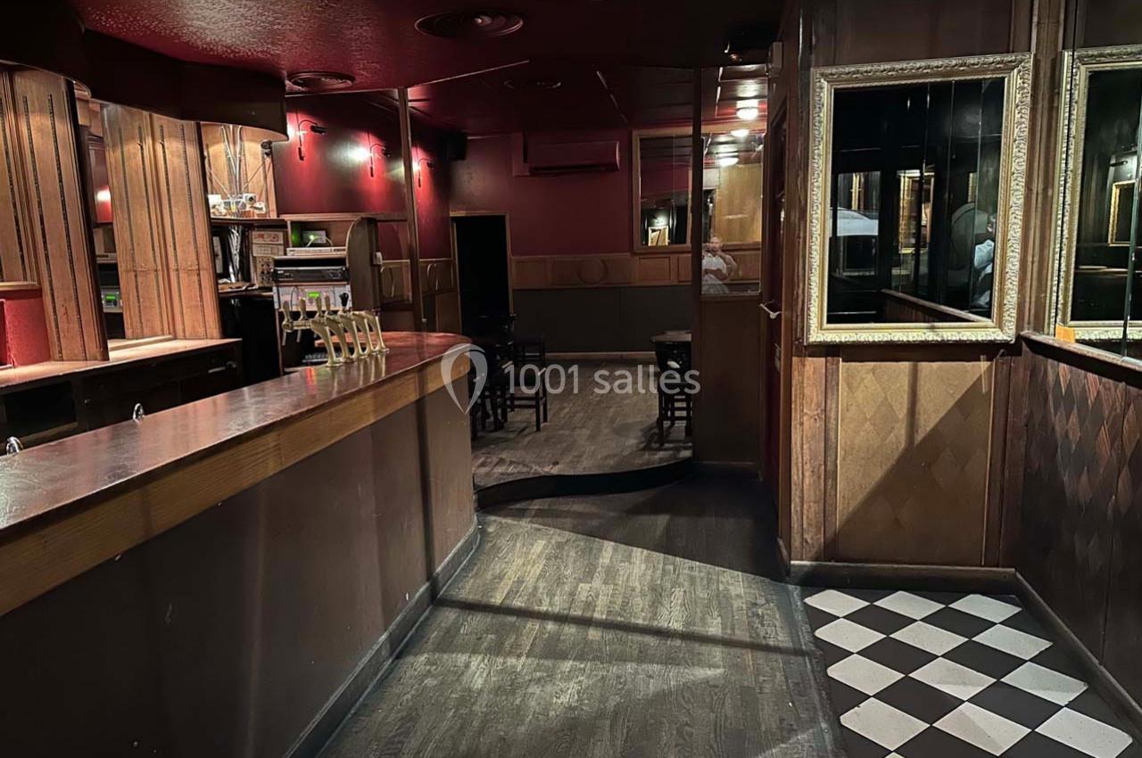 Intérieur d'un bar sombre avec comptoir en bois, sol en damier près de l'entrée et tables au fond de la pièce.