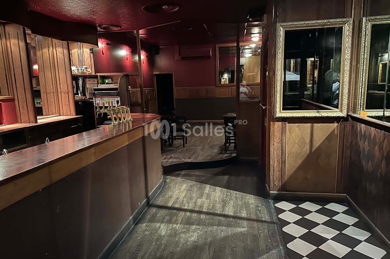 Intérieur d'un bar sombre avec comptoir en bois, sol en damier près de l'entrée et tables au fond de la pièce.