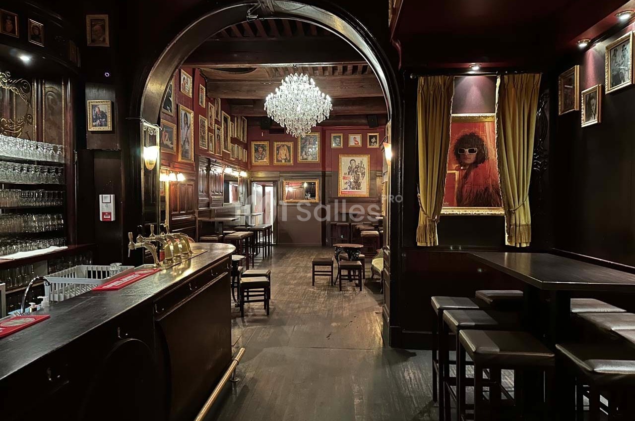 Intérieur d'un bar avec un comptoir en bois, des tables, un lustre central et des cadres accrochés aux murs rouges.