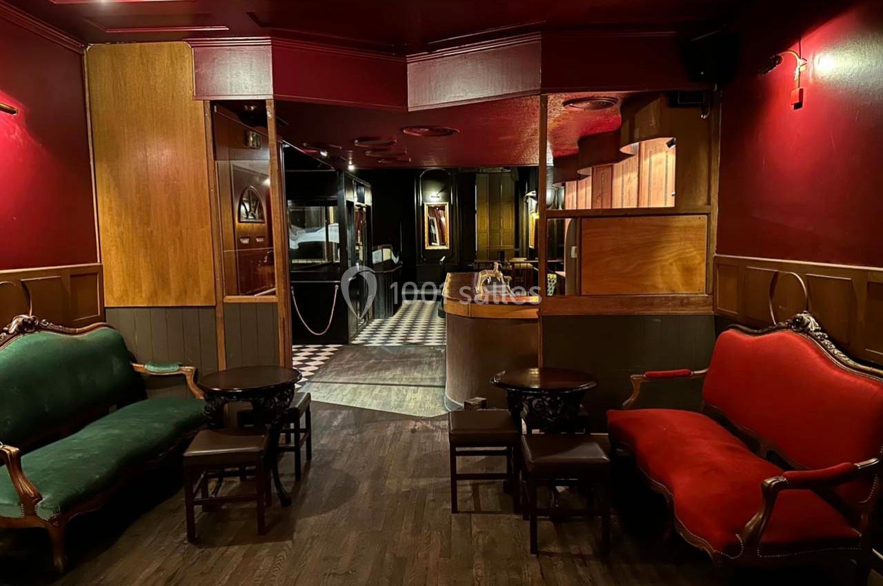 Intérieur d'un salon avec canapés rouges et verts, tables basses en bois, éclairage tamisé et sol en parquet.