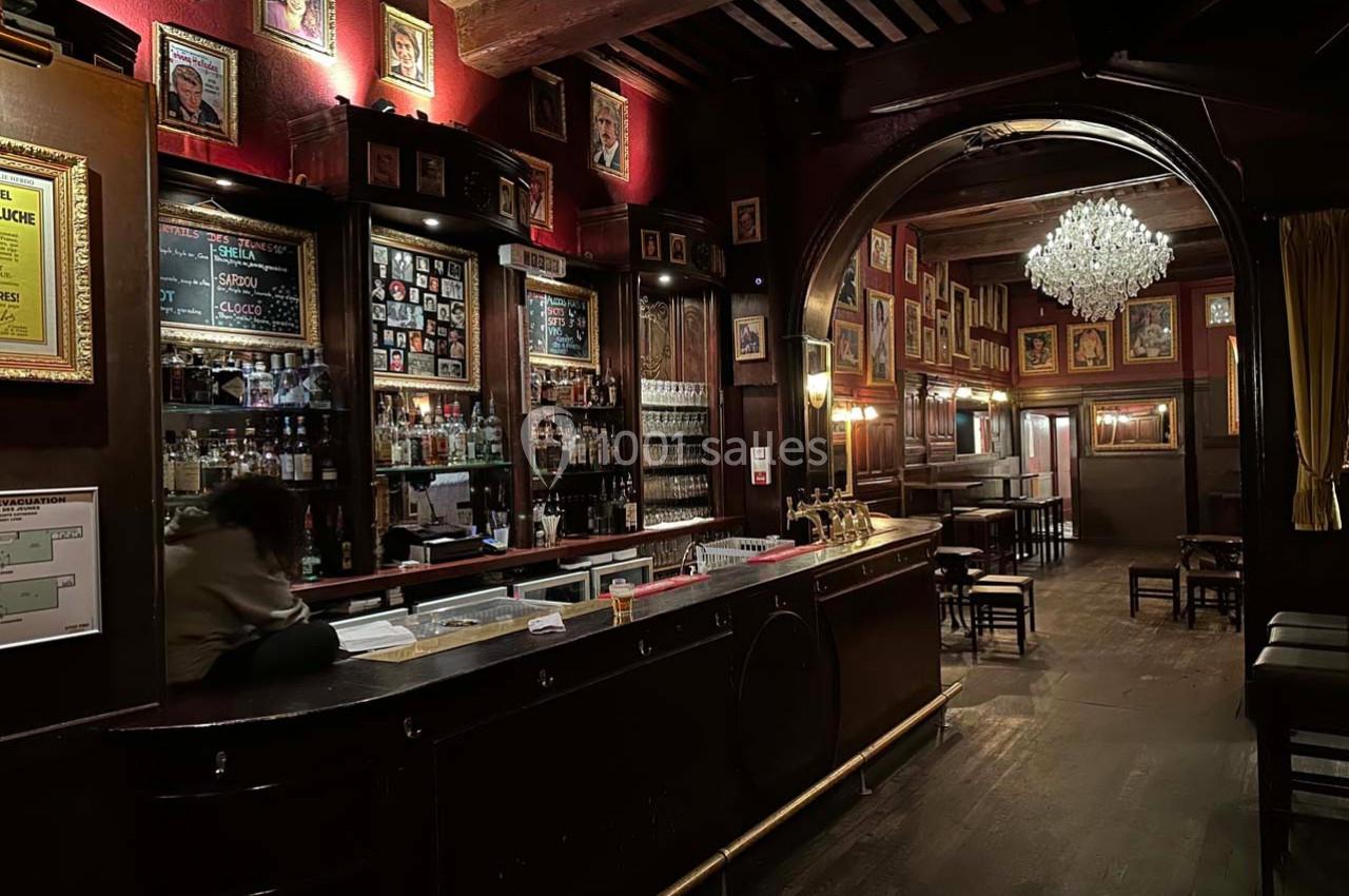 Intérieur d'un bar chaleureux avec un long comptoir, des étagères remplies de bouteilles et des murs décorés de cadres.