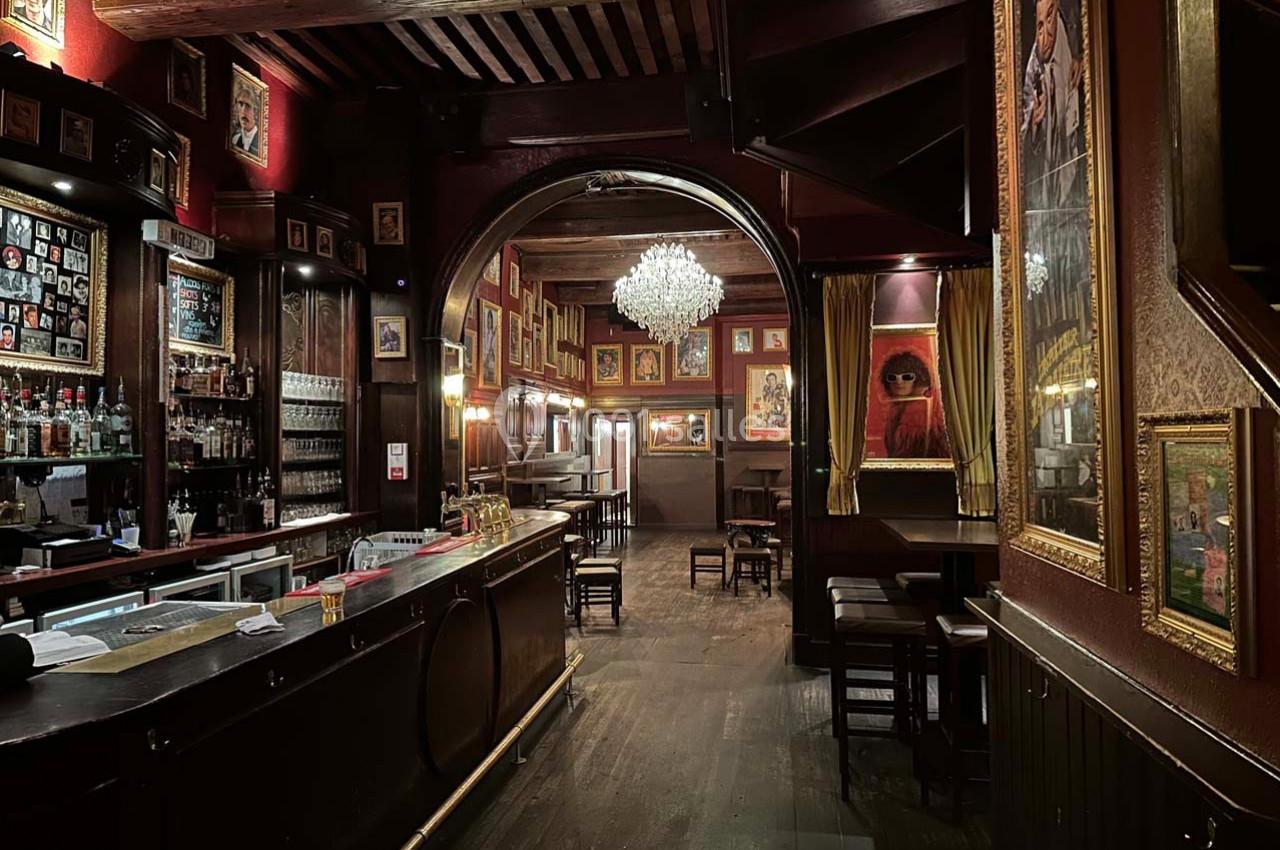 Intérieur d'un bar chaleureux avec un comptoir en bois, des tableaux aux murs et un lustre central éclairé.