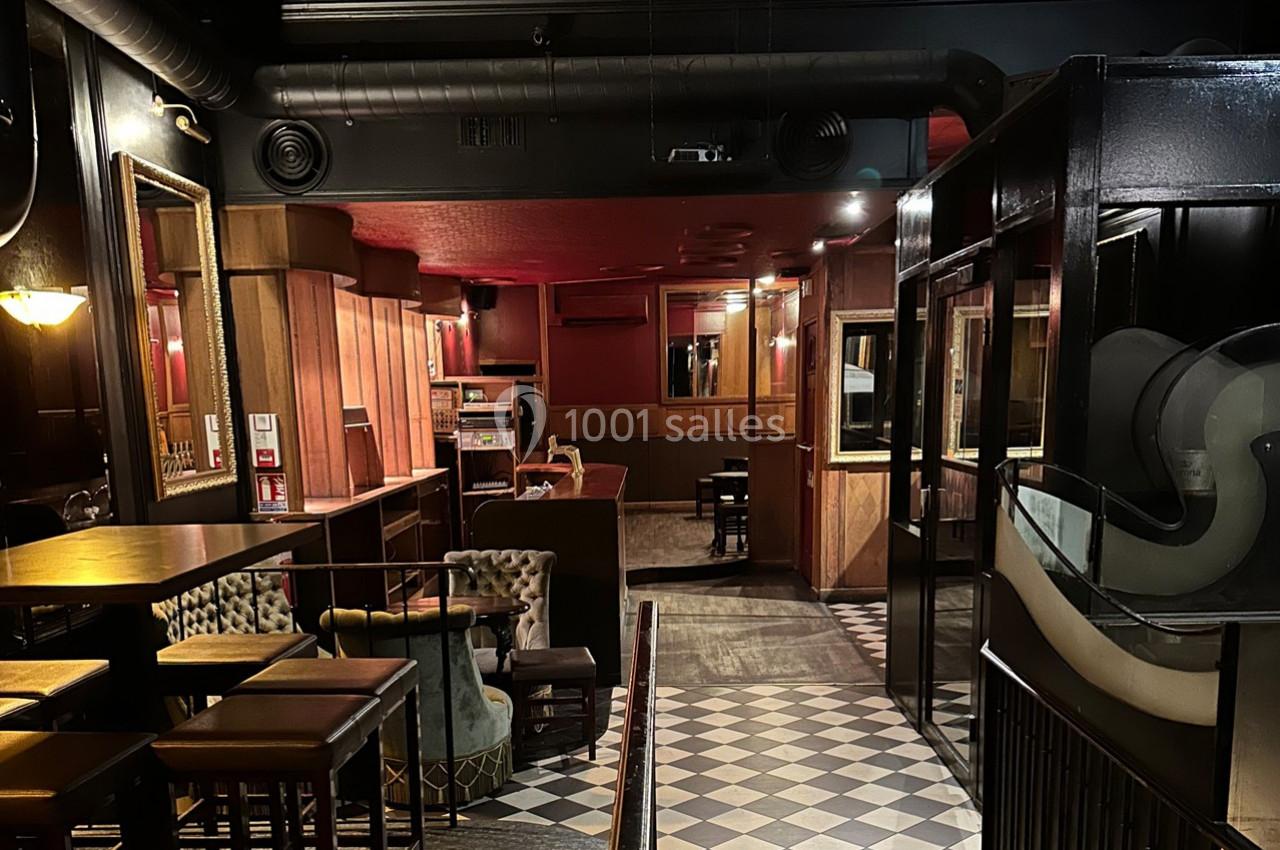 Intérieur d'un bar avec mobilier en bois, fauteuils, sol en damier et éclairage tamisé.